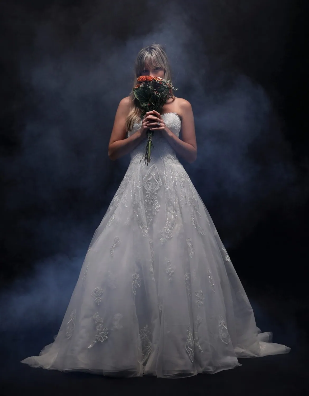 Bridal gown photographed on-model with a bouquet and smoke against a black backdrop